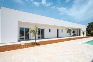 Un edificio moderno blanco con amplias puertas y ventanas de vidrio, una piscina y pequeñas palmeras al frente.