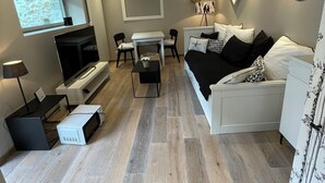 A modern living room with a flat-screen TV, a white sofa, a dining table with chairs, and a small kitchen area with a microwave.