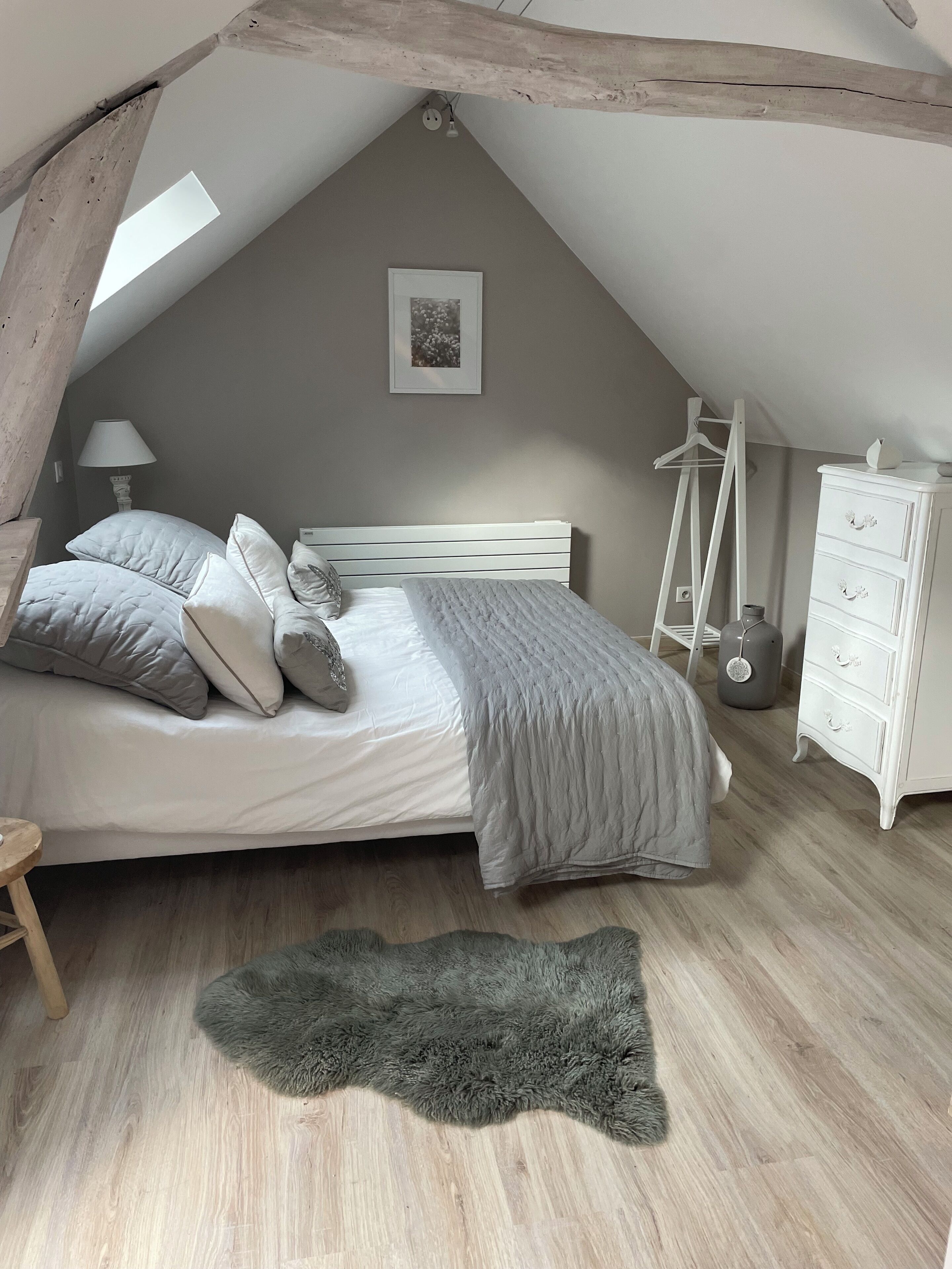 A bedroom with a sloped ceiling, a bed with grey bedding, a dresser, a lamp, and a hanging light fixture.