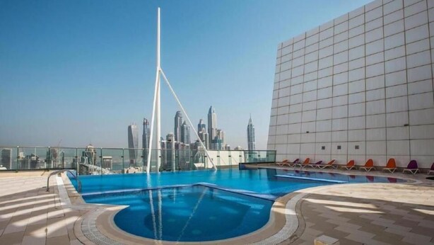 A rooftop pool with a city skyline view, a modern building with a grid facade, and a tall white structure. - Cosmos Living - Goldcrest Views1  (Dubai)