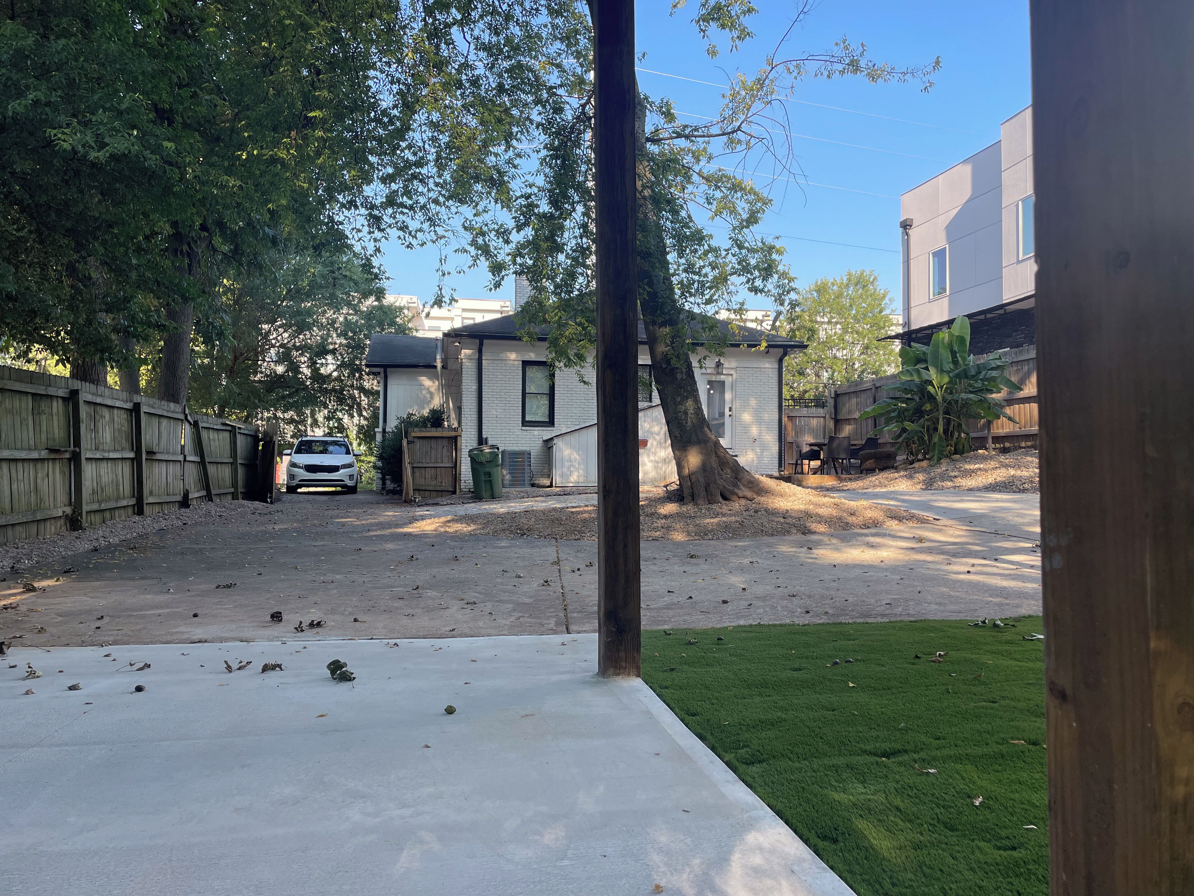 Un jardin à l’arrière d’une maison, avec une allée pavée, une clôture en bois et une voiture blanche garée au fond.