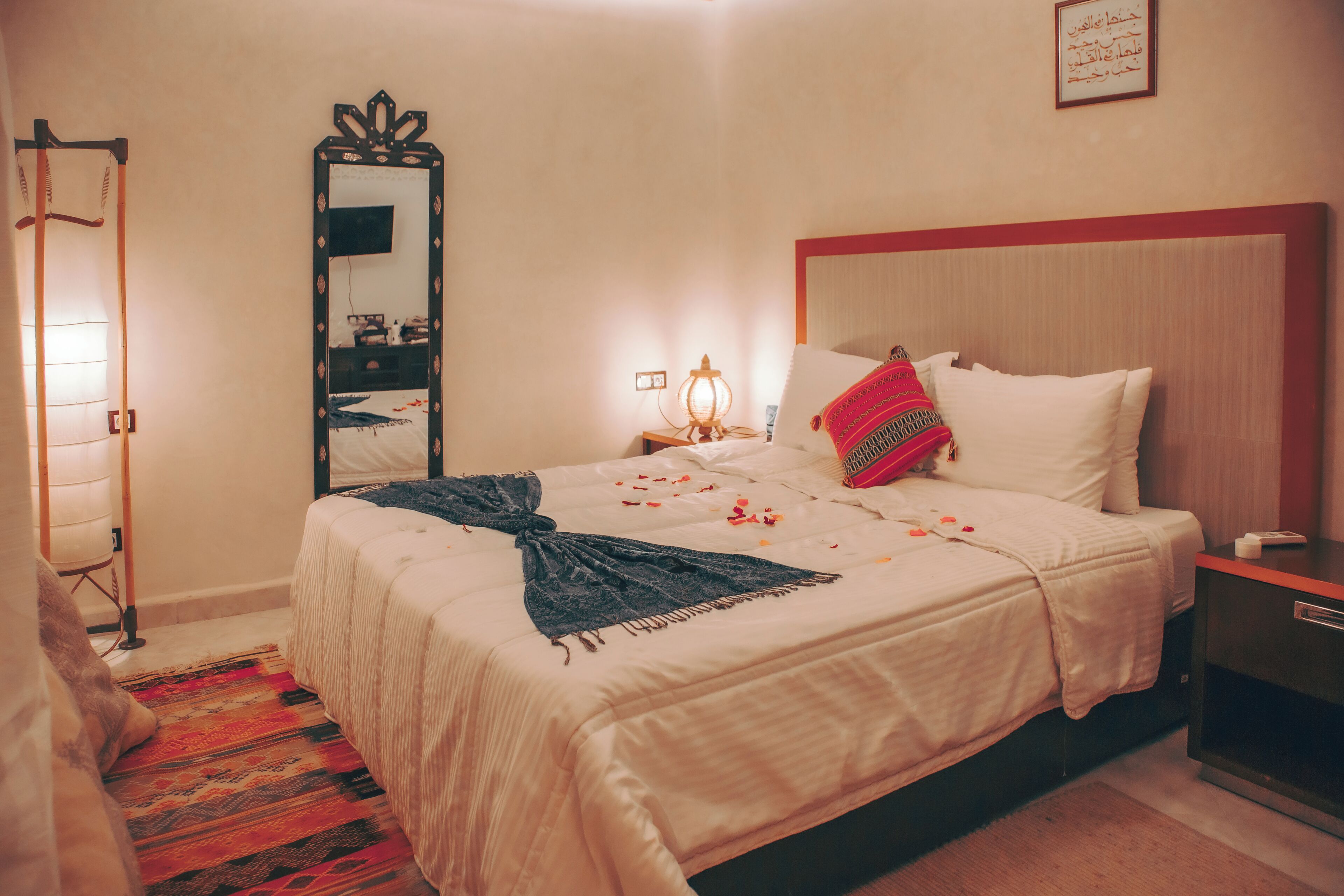 Un lit bien fait, recouvert d’une couette blanche, d’un plaid foncé et d’un coussin aux motifs rouges et noirs. Une table de chevet avec une lampe, un miroir et un tableau encadré sont posés sur le mur. Un tapis coloré recouvre le sol.
