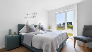 A modern bedroom with a bed, a nightstand, a chair, and a large window offering a view of the outdoors. - Malte's Ostseekoje- Infrared Sauna, Washing Machine, Terrace, Shower, Fireplace (Olpenitz)
