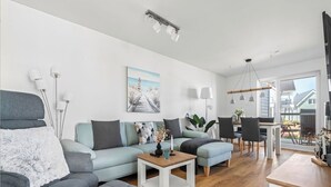 A modern living room with a grey sofa, a coffee table, and a dining area with chairs and a table. - Malte's Ostseekoje- Infrared Sauna, Washing Machine, Terrace, Shower, Fireplace (Olpenitz)