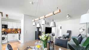 A modern kitchen with white cabinets, a built-in oven, and a microwave. There is a dining area with a wooden table, chairs, and a vase of flowers. - Malte's Ostseekoje- Infrared Sauna, Washing Machine, Terrace, Shower, Fireplace (Olpenitz)