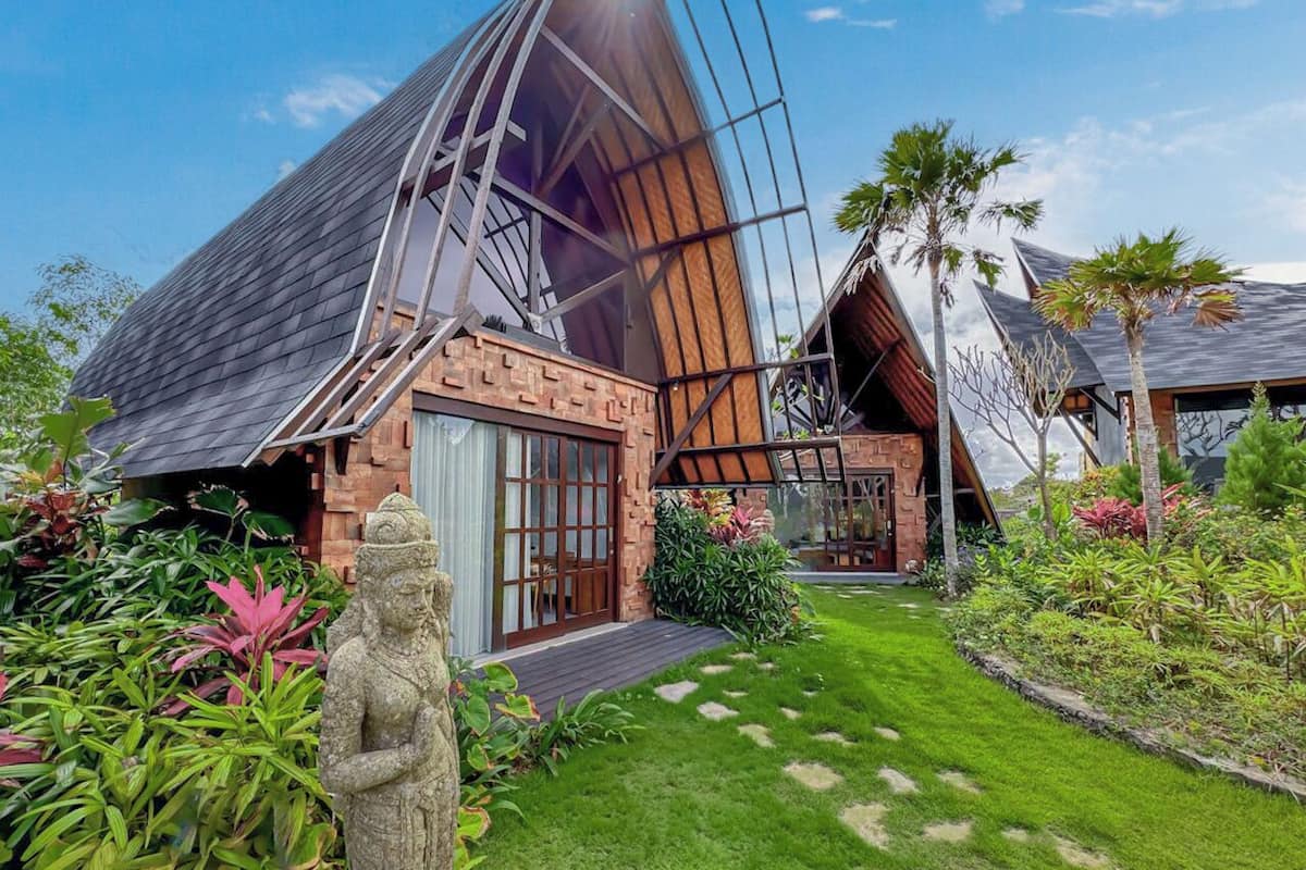 A modern house with a unique curved roof, surrounded by lush greenery and a statue.