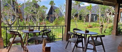 A covered outdoor seating area with wooden tables and chairs, a decorative hanging light, and a view of a well-maintained garden and buildings in the background.