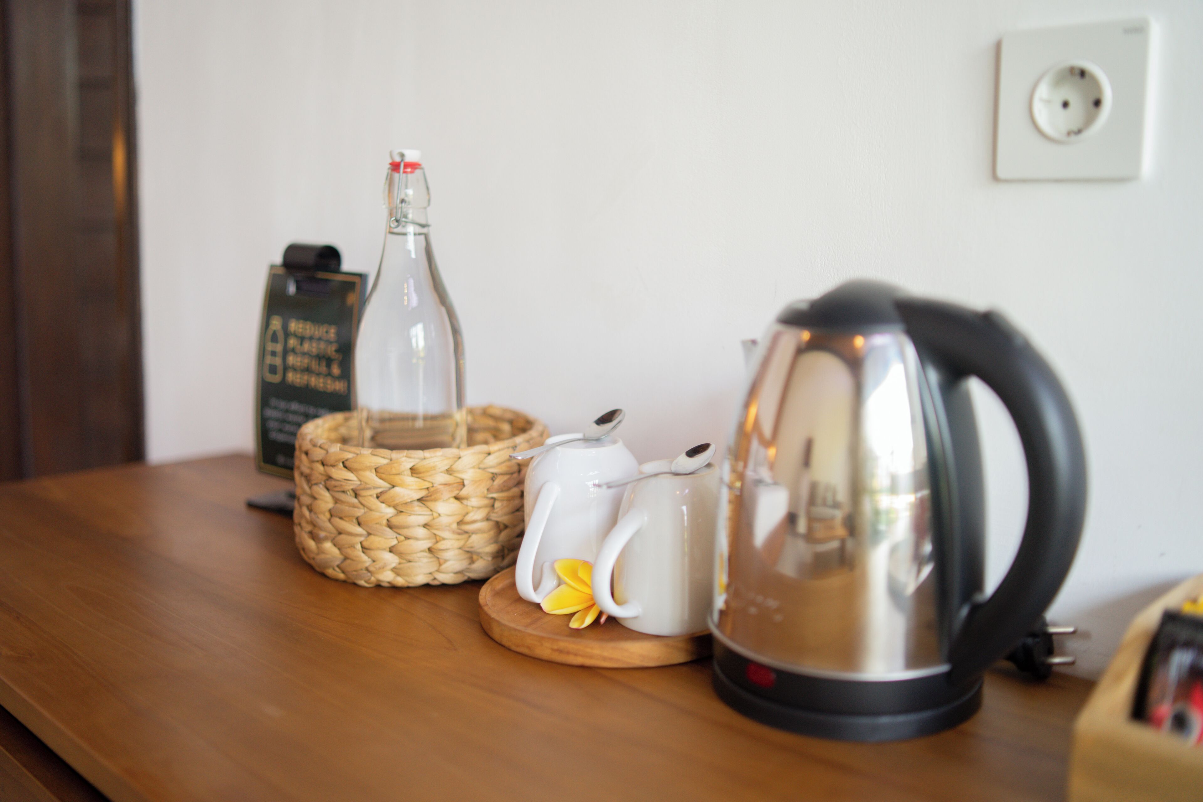 A wooden table with a woven basket, a glass bottle, a white teapot, and a black electric kettle.
