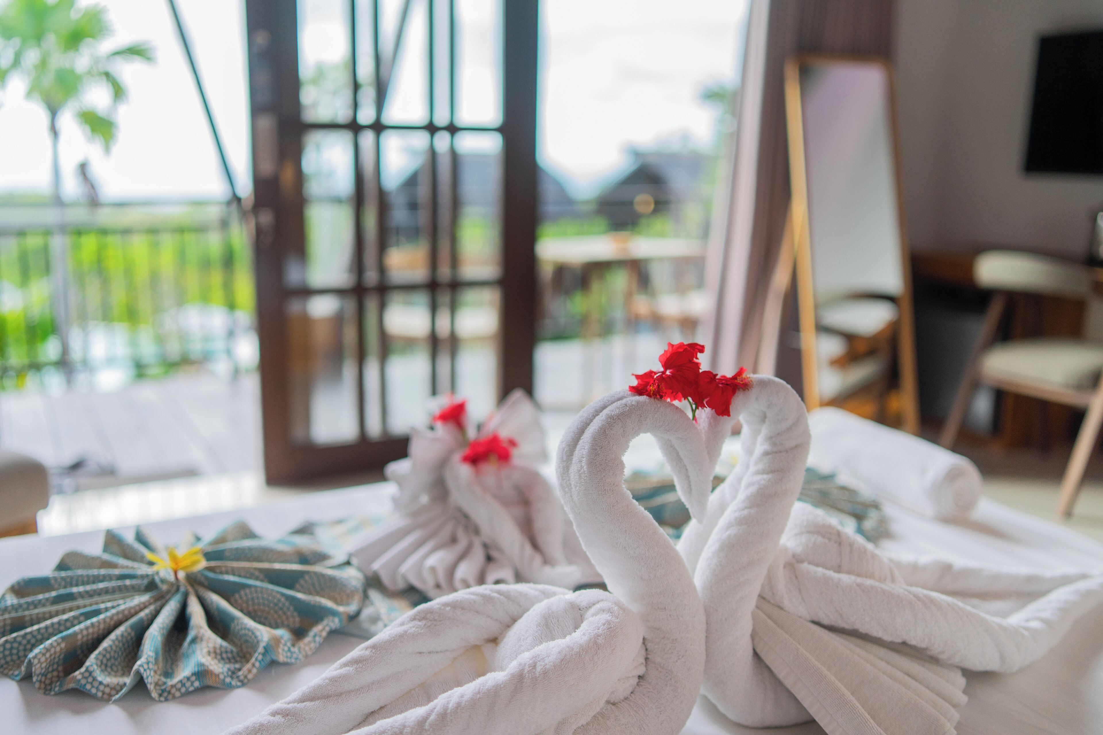 A room with a view of a balcony, a table with towels arranged in swan shapes, and a patterned cloth on the table.