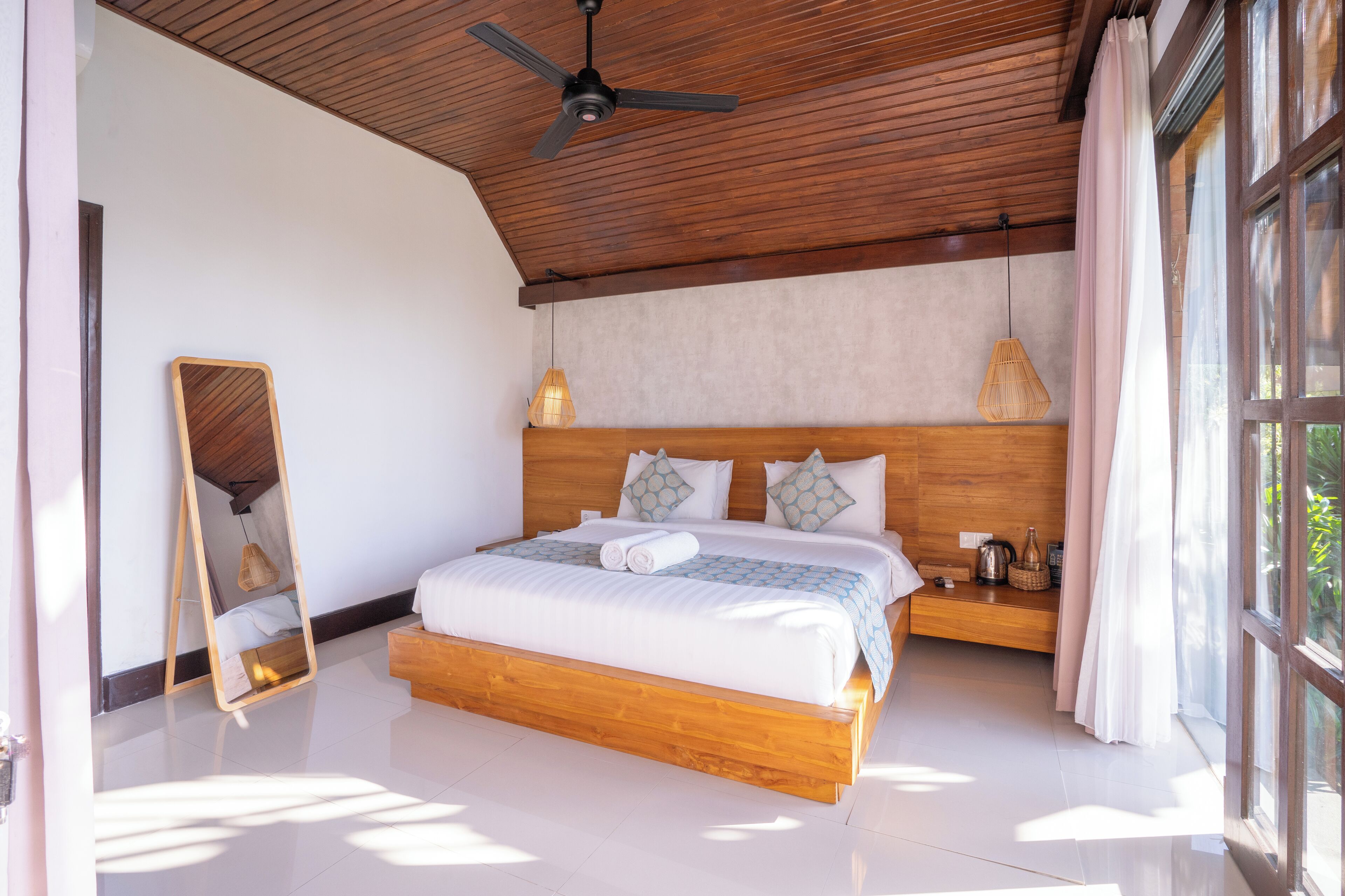 Un dormitorio con estructura de cama de madera, ventilador de techo y un amplio ventanal con cortinas.