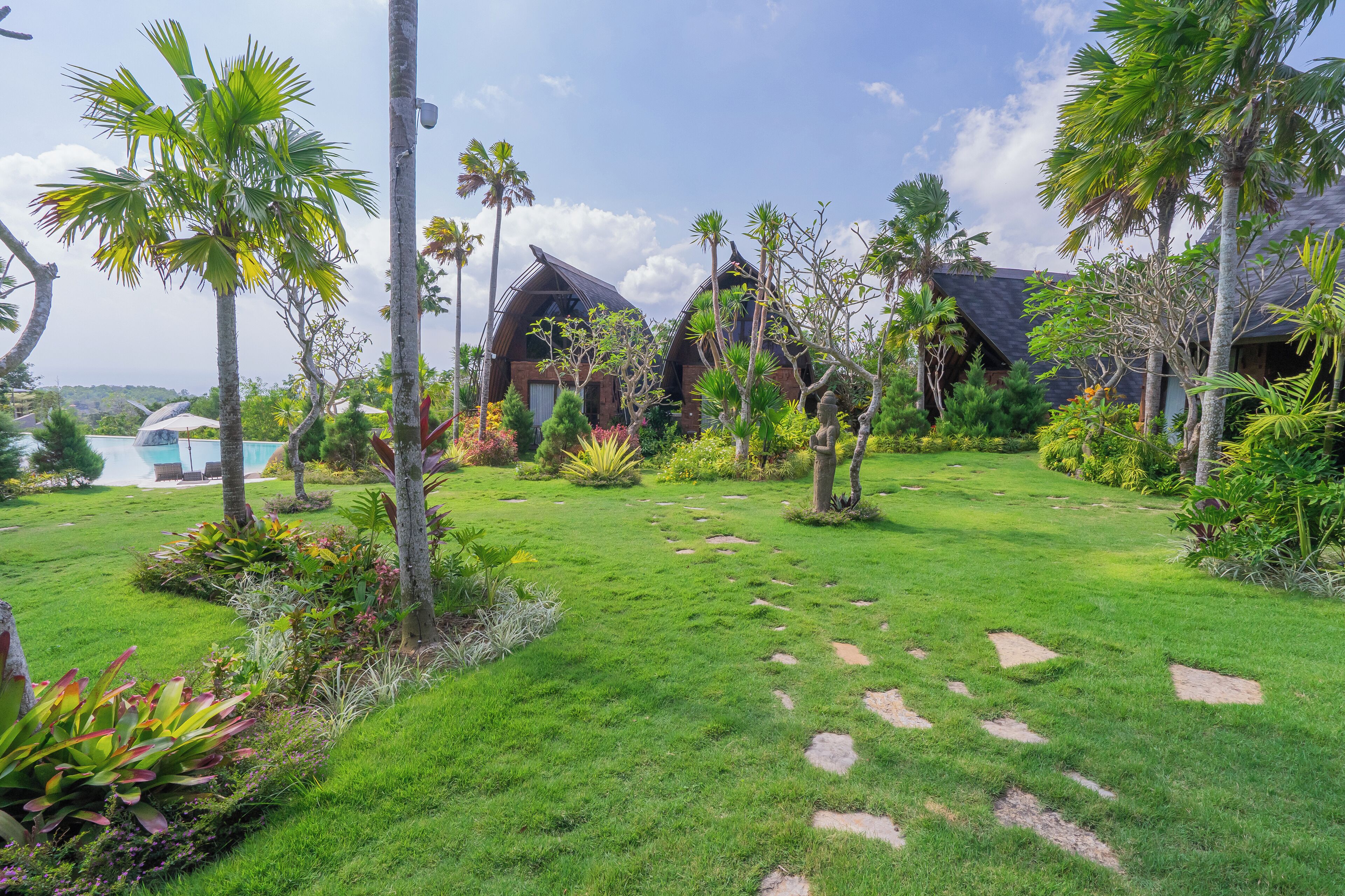 A lush garden with a pool, palm trees, and a building with a sloped roof.