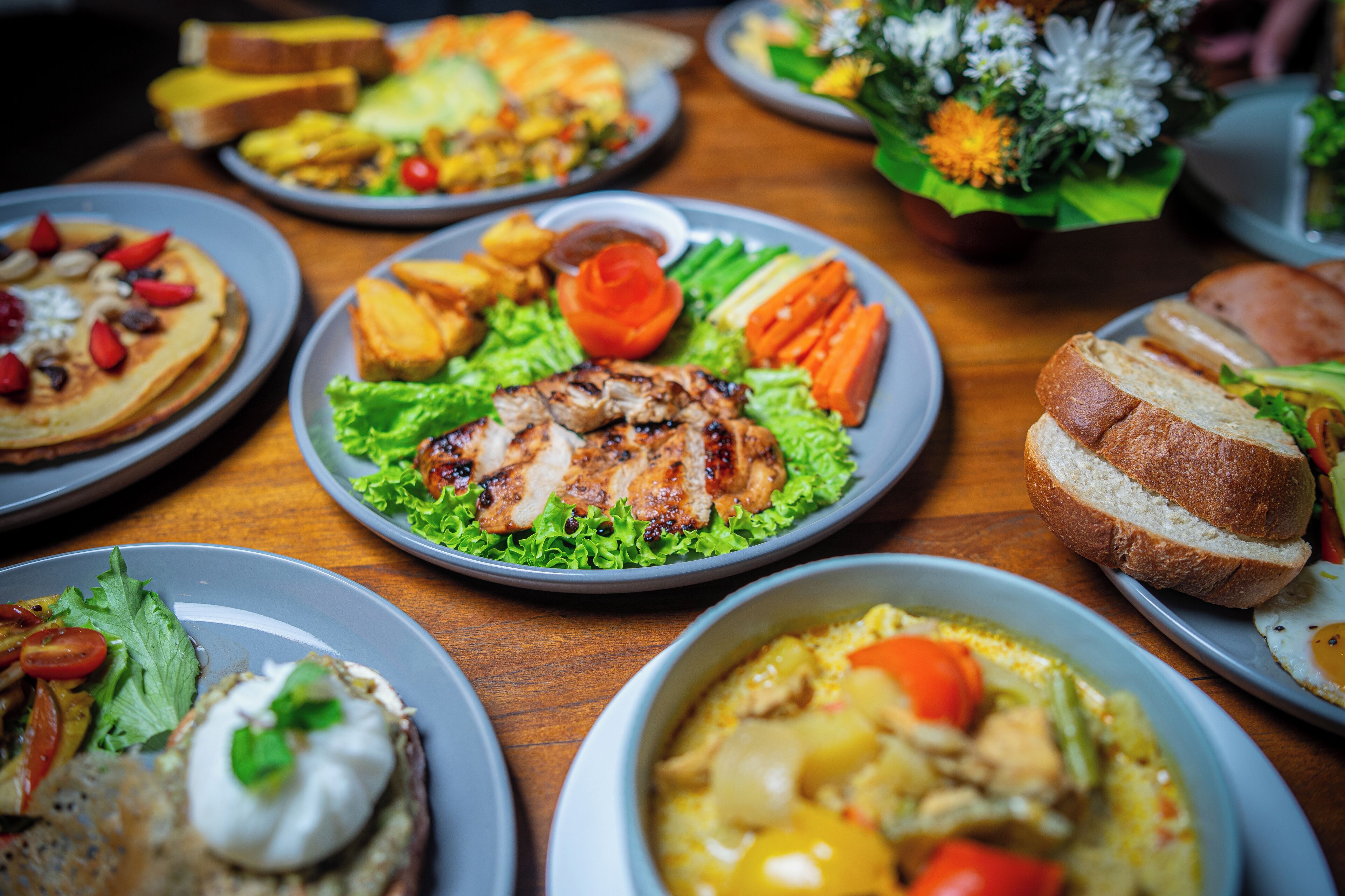 A variety of dishes on a wooden table, including grilled chicken, vegetables, and bread.