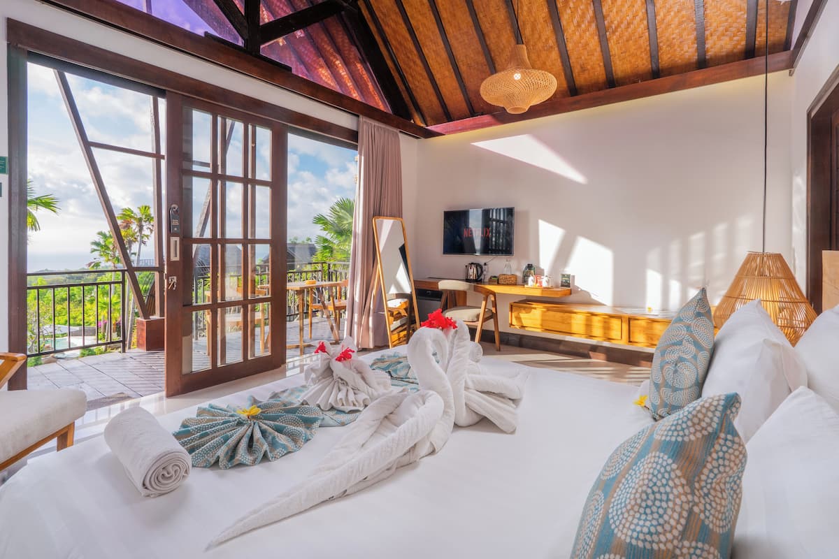 A hotel room with a bed, a desk, and a TV. There is a balcony with a view of palm trees and the ocean.