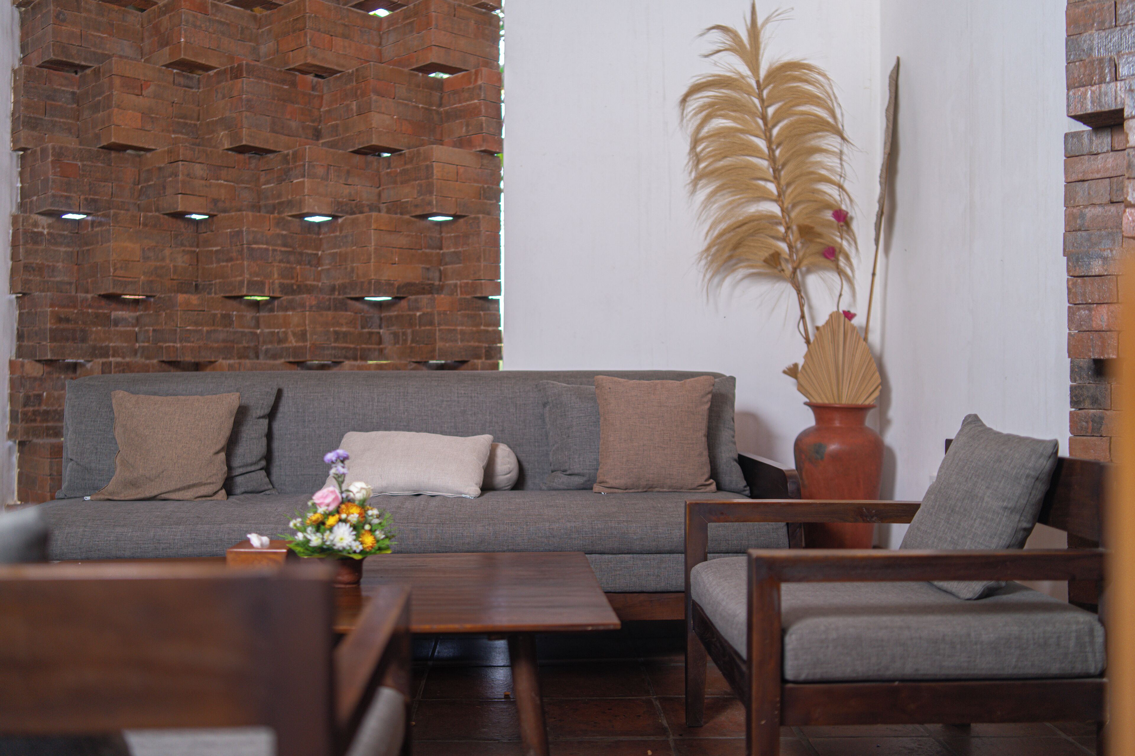 Ein Wohnzimmer mit grauem Sofa, Holz-Couchtisch und einem Sessel. Eine Ziegelwand weist ein geometrisches Muster auf, und eine Vase enthält getrocknete Pflanzen.