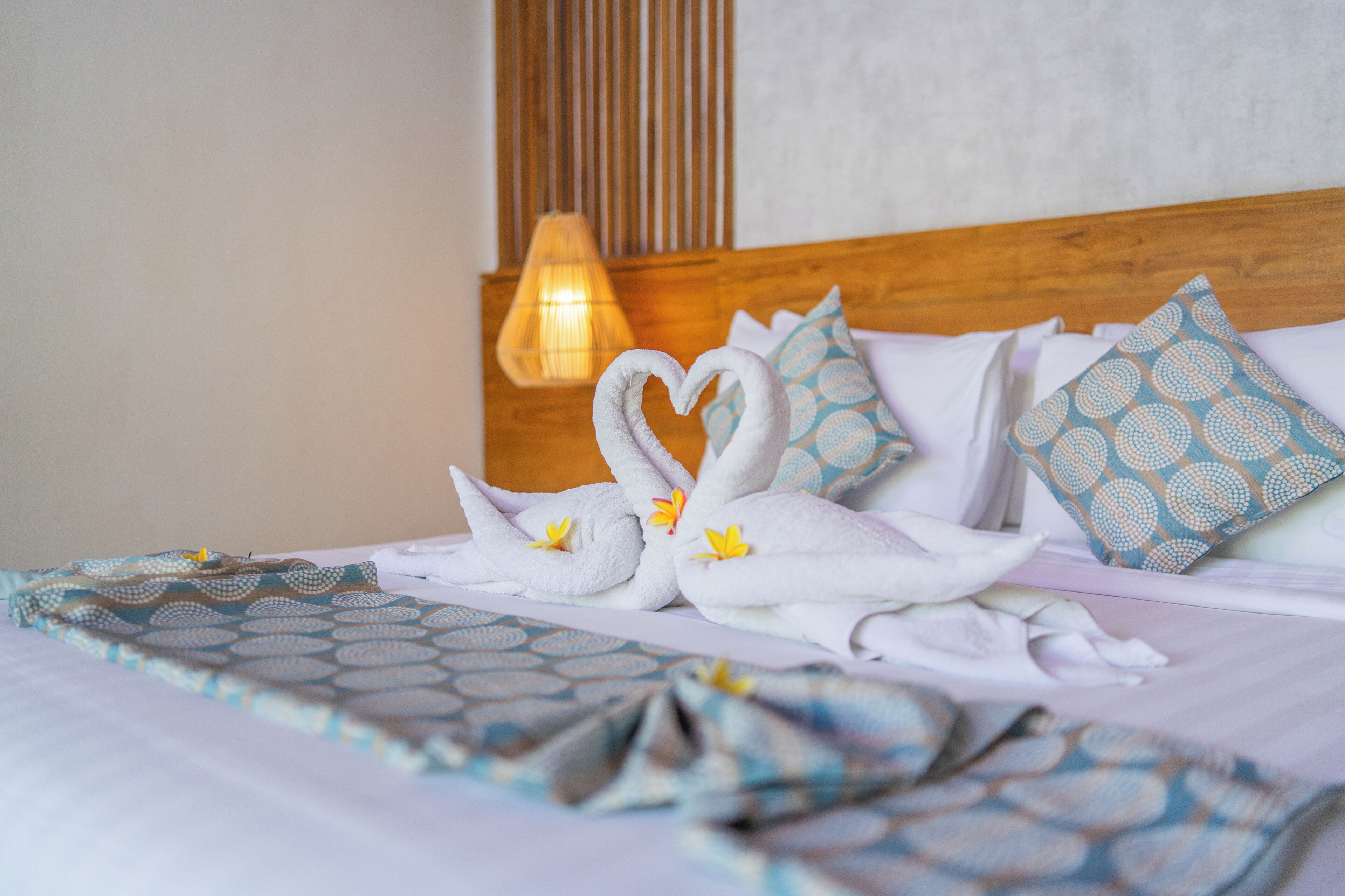 Una cama impecablemente arreglada con una toalla dispuesta en forma de corazón, almohadas estampadas y una cálida iluminación ambiental.