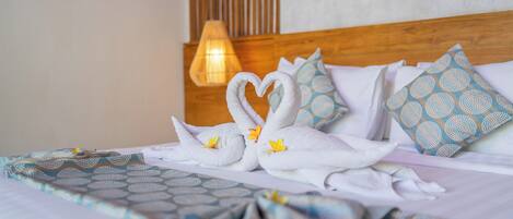 Una cama impecablemente arreglada con una toalla dispuesta en forma de corazón, almohadas estampadas y una cálida iluminación ambiental.
