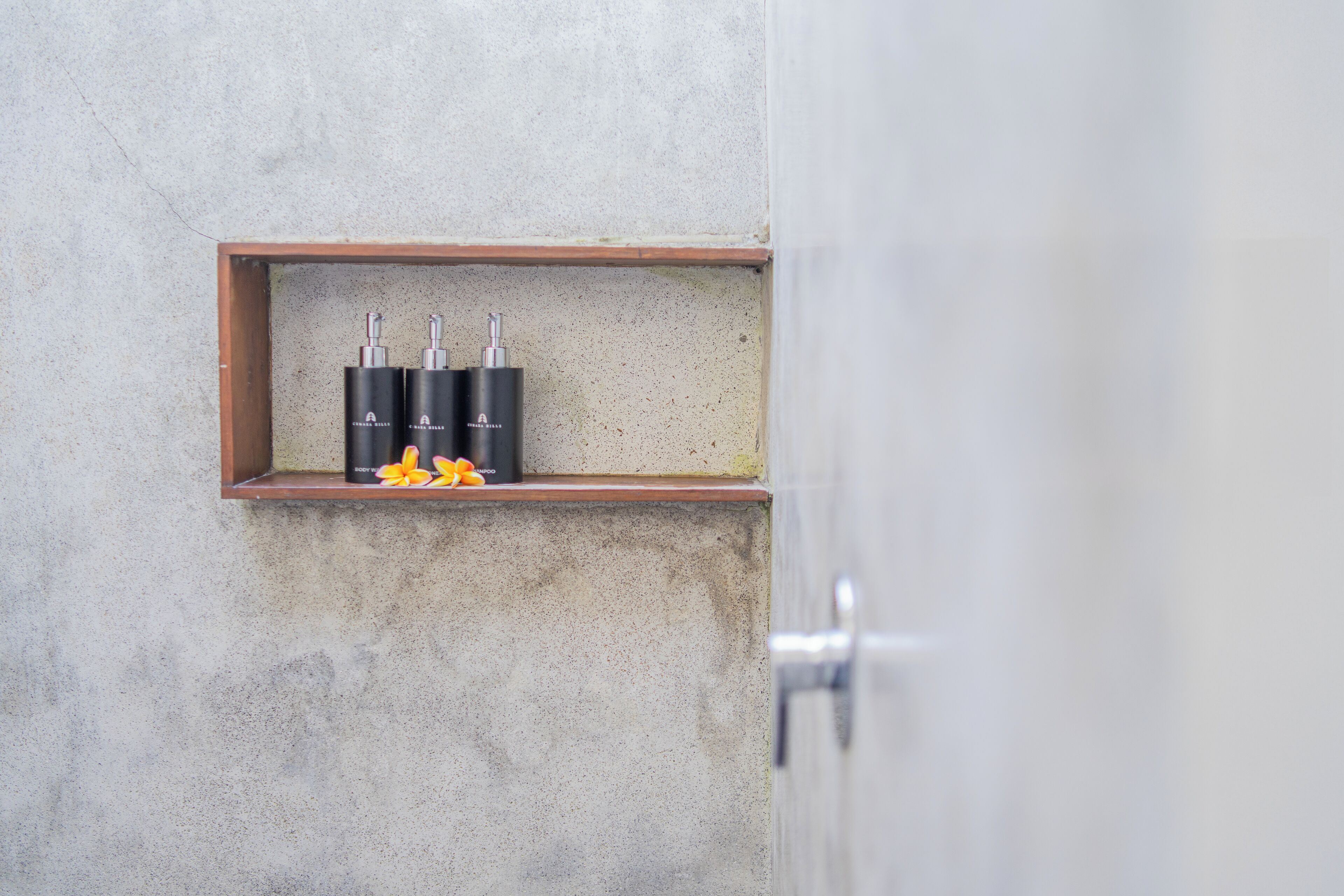 A bathroom with a wooden shelf holding three black bottles and a few yellow flowers.