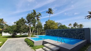 A rectangular swimming pool with a stone wall and metal ladder, surrounded by a paved walkway and lush greenery.