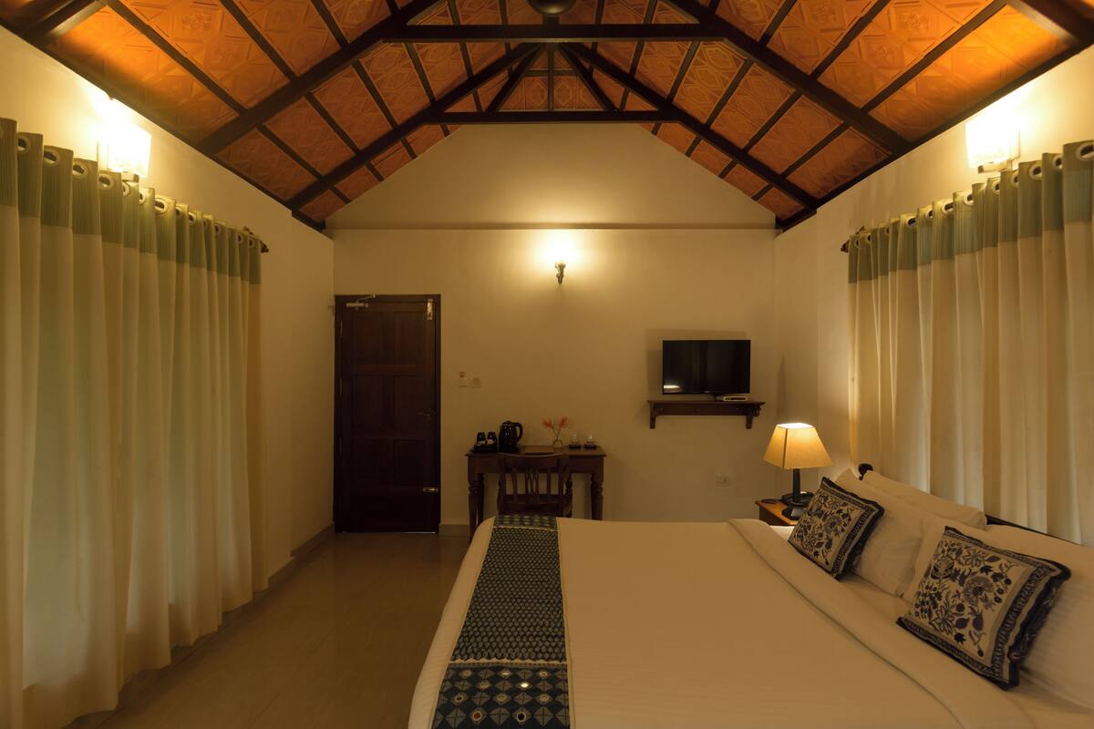 Une chambre d’hôtel avec un plafond en bois, un lit agrémenté d’oreillers à motifs, une table de chevet avec une lampe et un téléviseur fixé au mur.