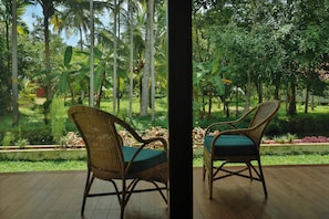 Two wicker chairs with green cushions on a wooden deck, overlooking a lush garden with various plants and trees. - Palmgrove Lake Resort (Ambalapuzha)