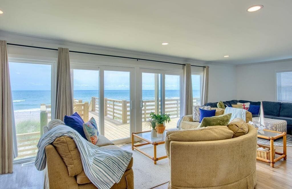 Un salón amplio con un gran ventanal con vistas a la playa, amueblado con un sofá, sillones y una mesa de centro.