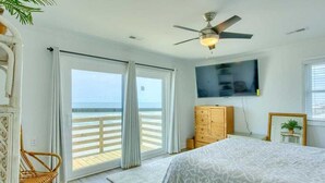 A bedroom with a bed, a television, a ceiling fan, a wicker chair, and a view of the beach through the sliding doors. - The Black Pearl - Oceanfront - North Topsail (North Topsail Beach)