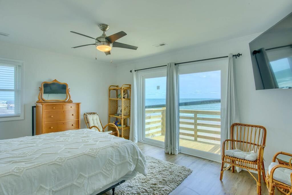 A bedroom with a bed, a dresser, a chair, a bookshelf, and a view of the ocean through a sliding door.