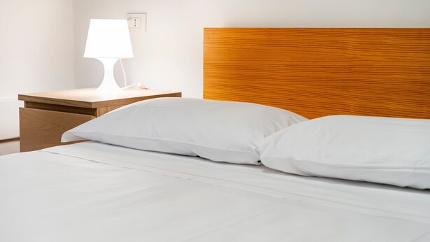 Una cama bien hecha con sábanas y almohadas blancas, un cabecero de madera y una mesita de noche con una lámpara blanca.