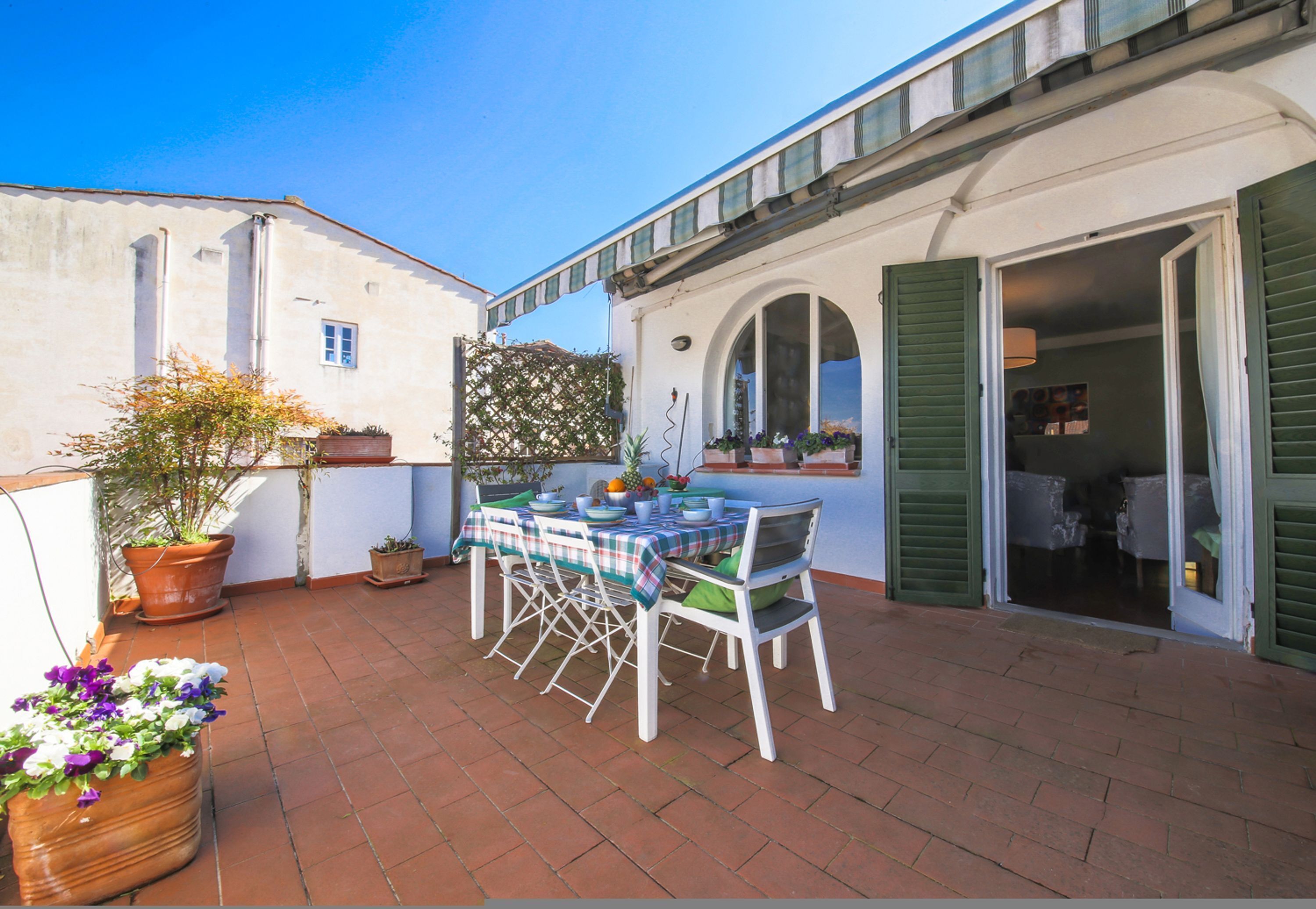 A patio with a table and chairs, potted plants, and a building with green shutters.