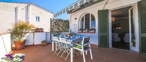 A patio with a table and chairs, potted plants, and a building with green shutters.