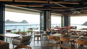 Un restaurant moderne offrant une vue sur la mer, avec des tables et des chaises en bois, de grandes fenêtres et un design contemporain au niveau du plafond.