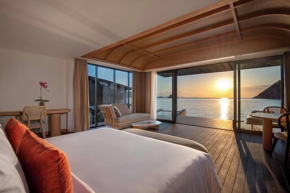 Ein modernes Hotelzimmer mit einem großen Bett, einem Schreibtisch und einem Balkon mit Blick auf den Sonnenuntergang über dem Wasser.