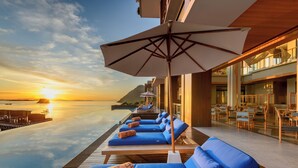 Une piscine à débordement baignée de soleil, dotée de chaises longues et de parasols, qui offre une vue sur une étendue d’eau et une île lointaine.