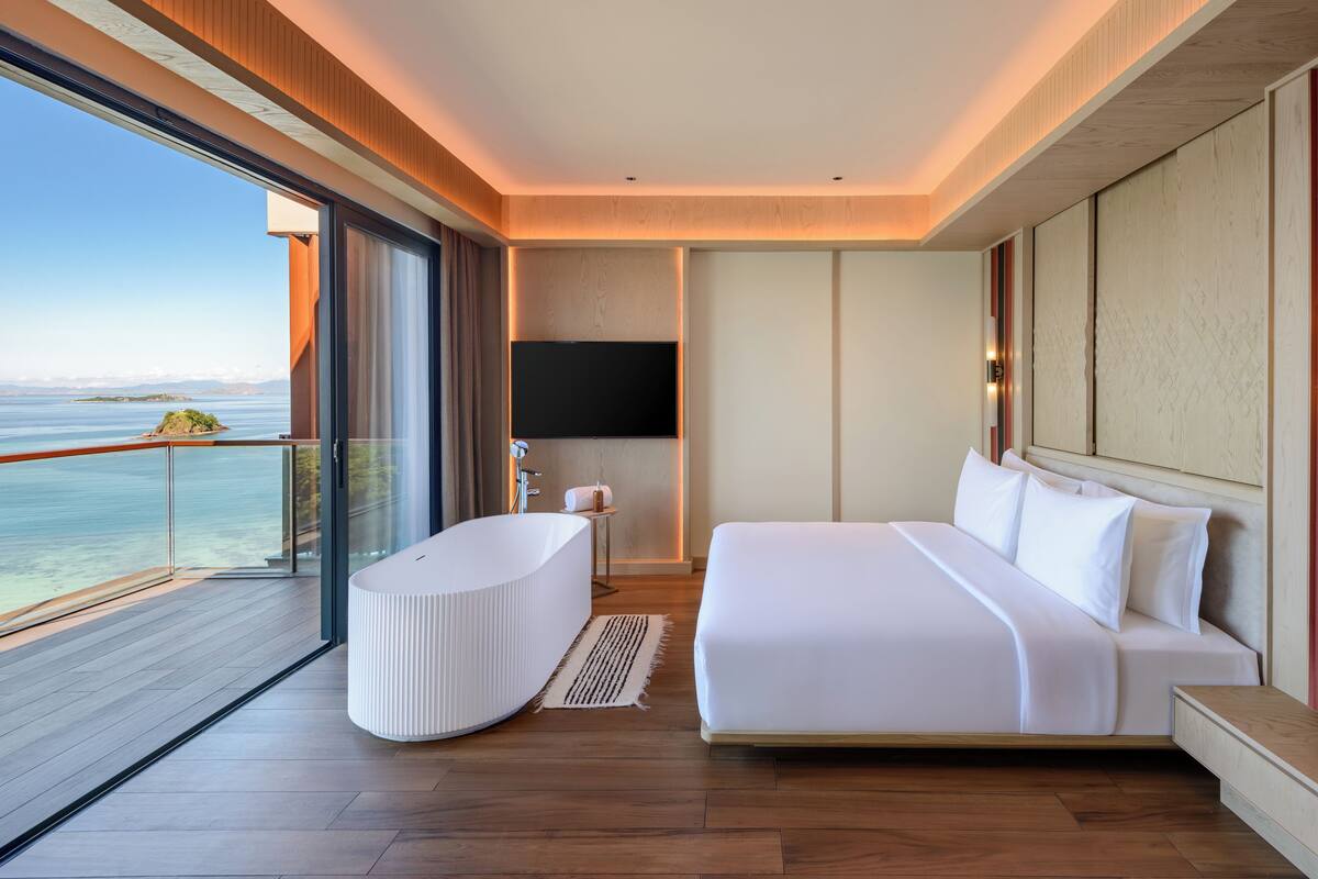 Ein modernes Hotelzimmer mit einem großen Bett, einer Badewanne, einem Fernseher und einem Balkon mit Meerblick.