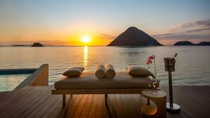 Une terrasse en bois avec un fauteuil de massage et une petite table sur laquelle se trouvent des verres et une bouteille, donnant sur une mer calme au coucher du soleil.