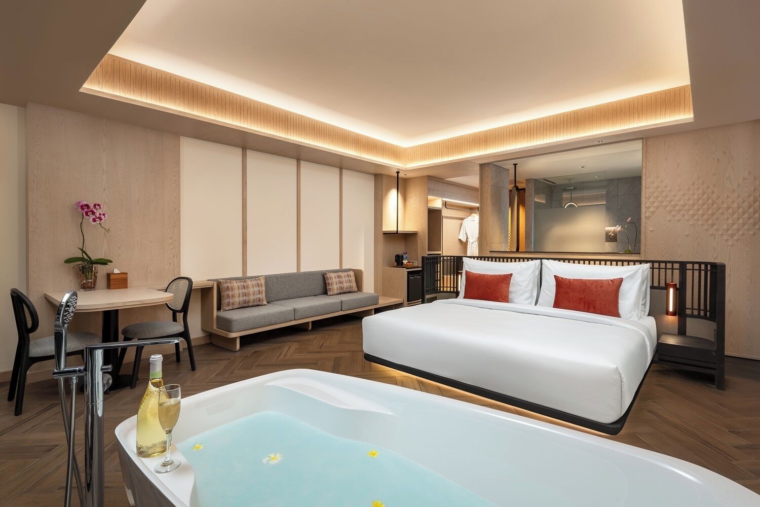 A modern hotel room with a large bed, a sitting area with a sofa, a dining table, and a separate bathroom with a bathtub.