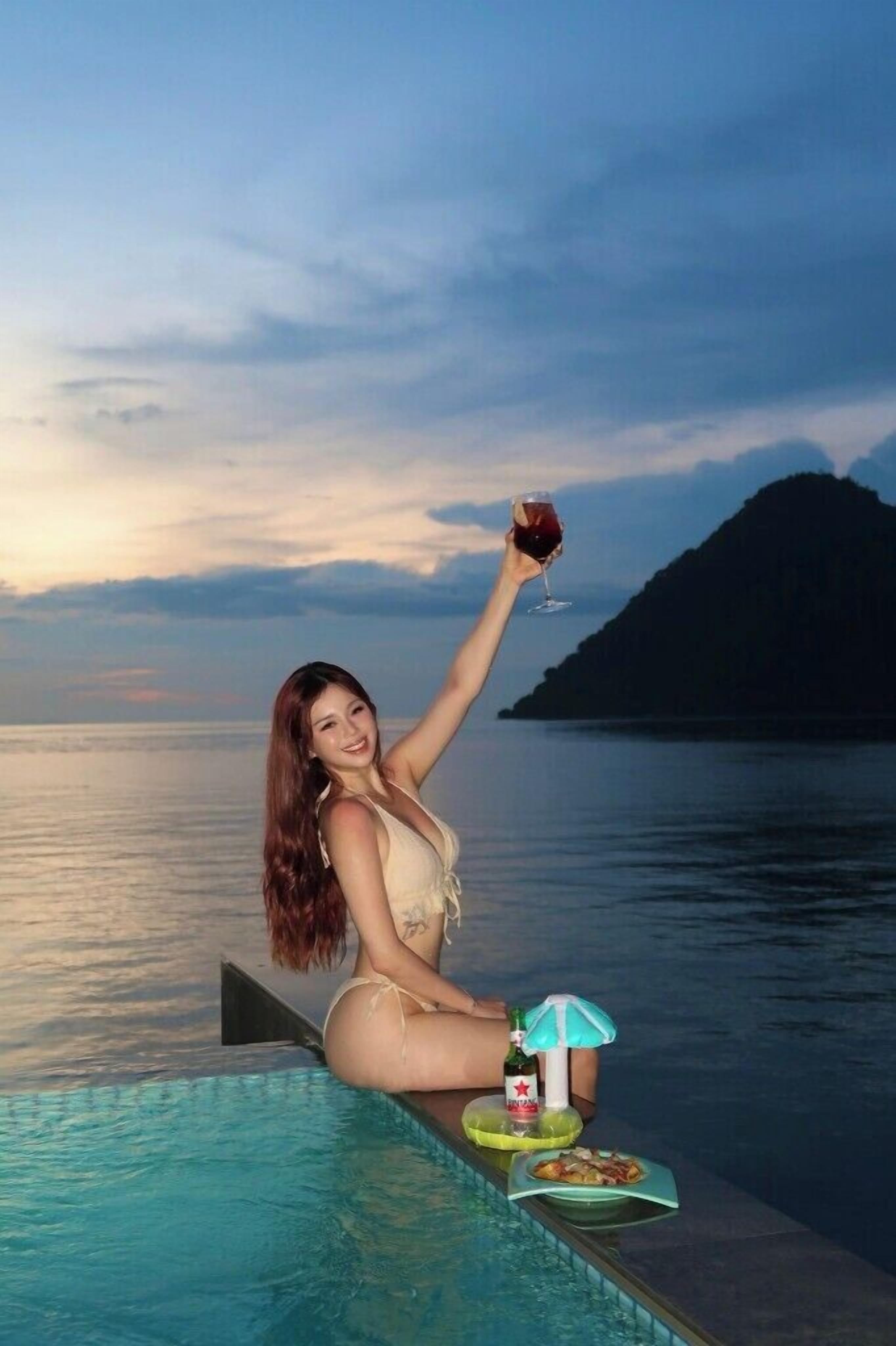 A person sitting by a pool with a drink, a bottle, and a plate of food.
