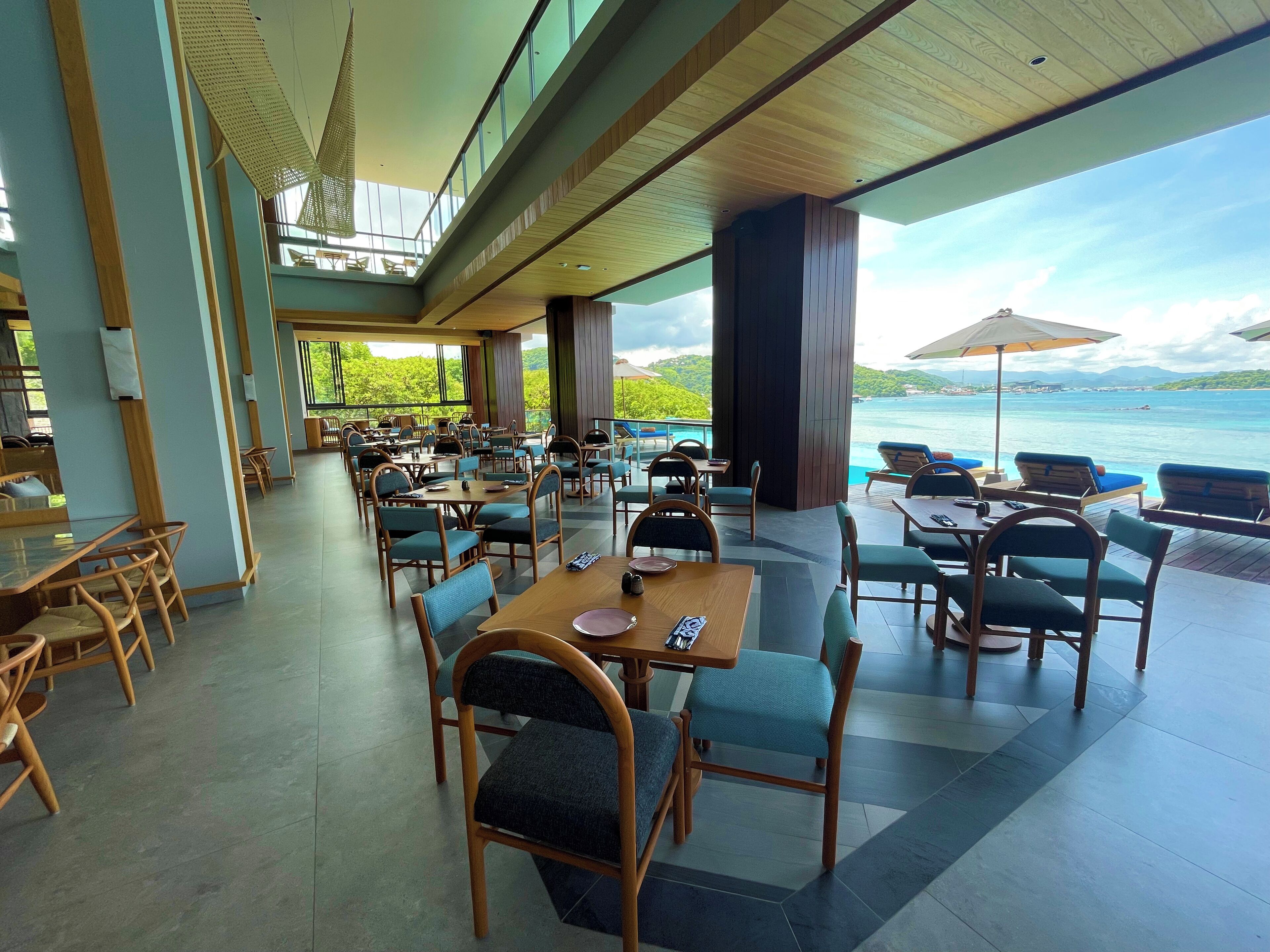 A spacious outdoor dining area with wooden tables and chairs, large windows offering a view of the sea and mountains.