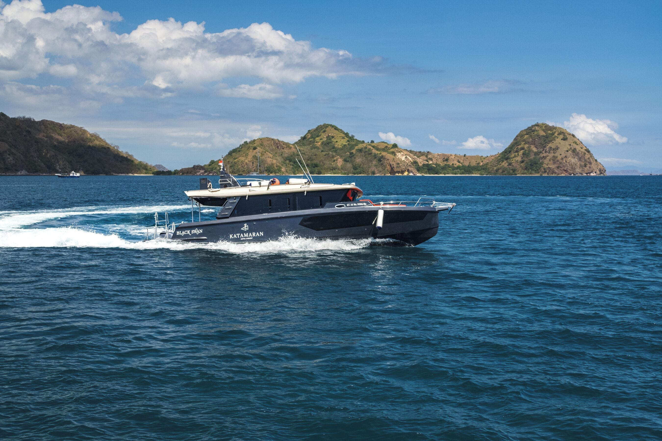 A boat named 'Black Swan' with 'Katamaran' written on the side, moving on a body of water with hills in the background.