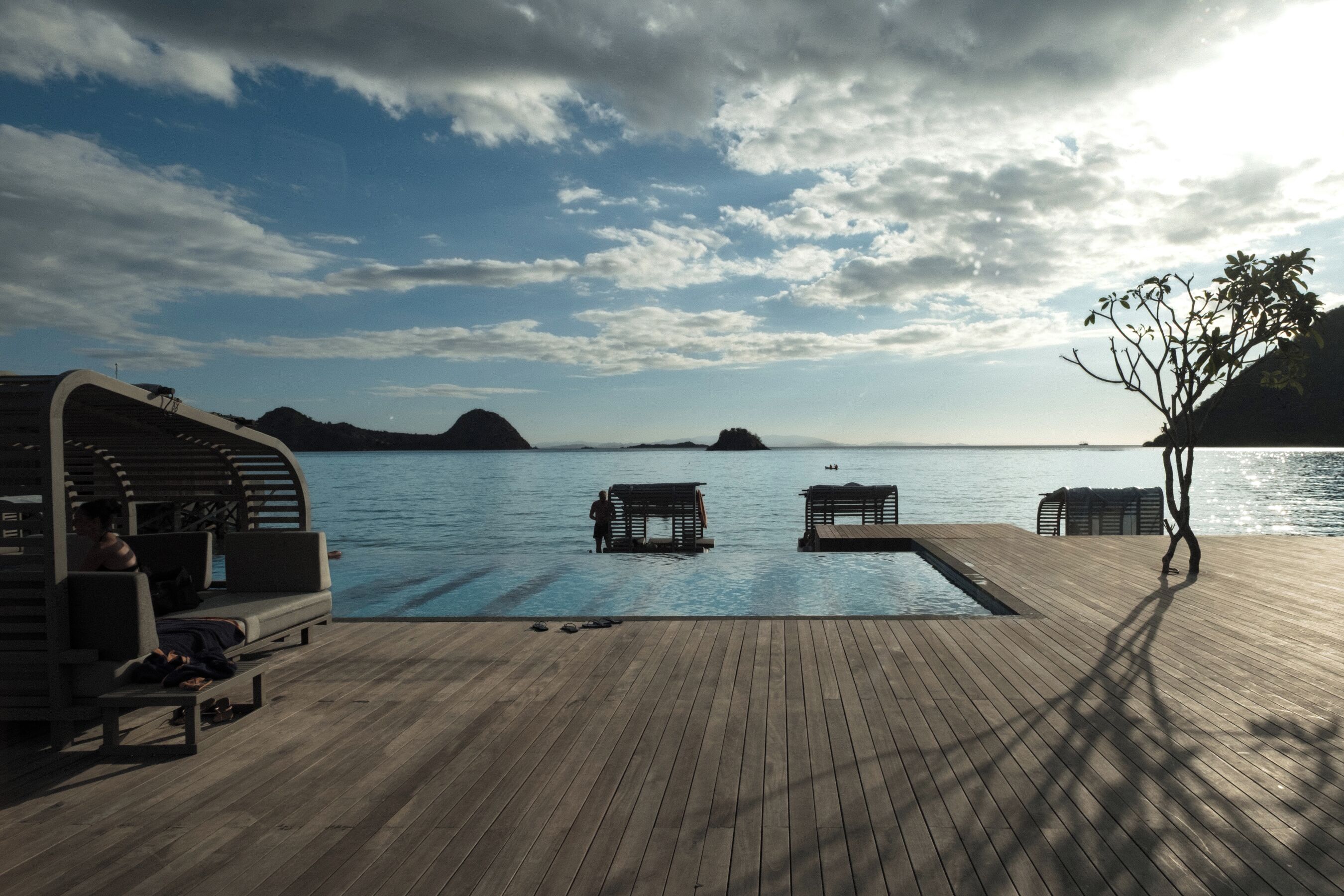 A wooden deck with lounge chairs and a pool, overlooking calm water and distant islands.