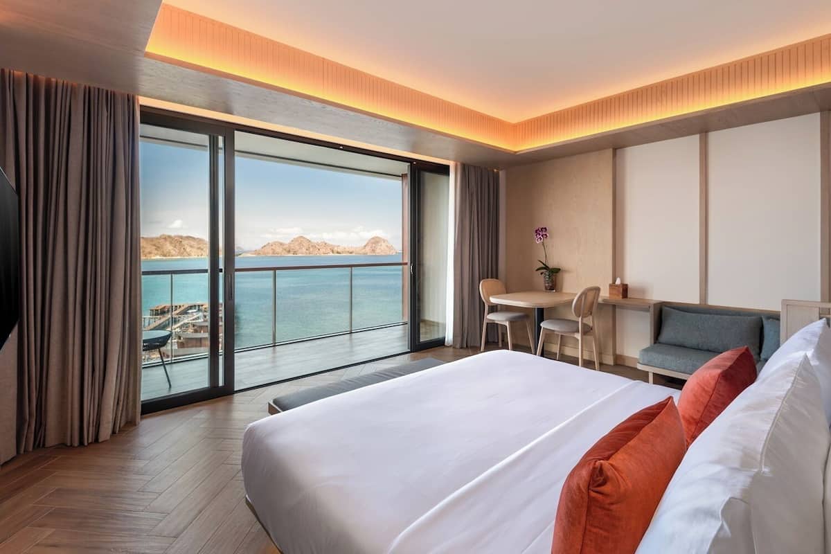 Ein modernes Hotelzimmer mit einem großen Bett, einem Schreibtisch, einer Sitzecke und einem Balkon mit Blick auf das Meer und die Berge.