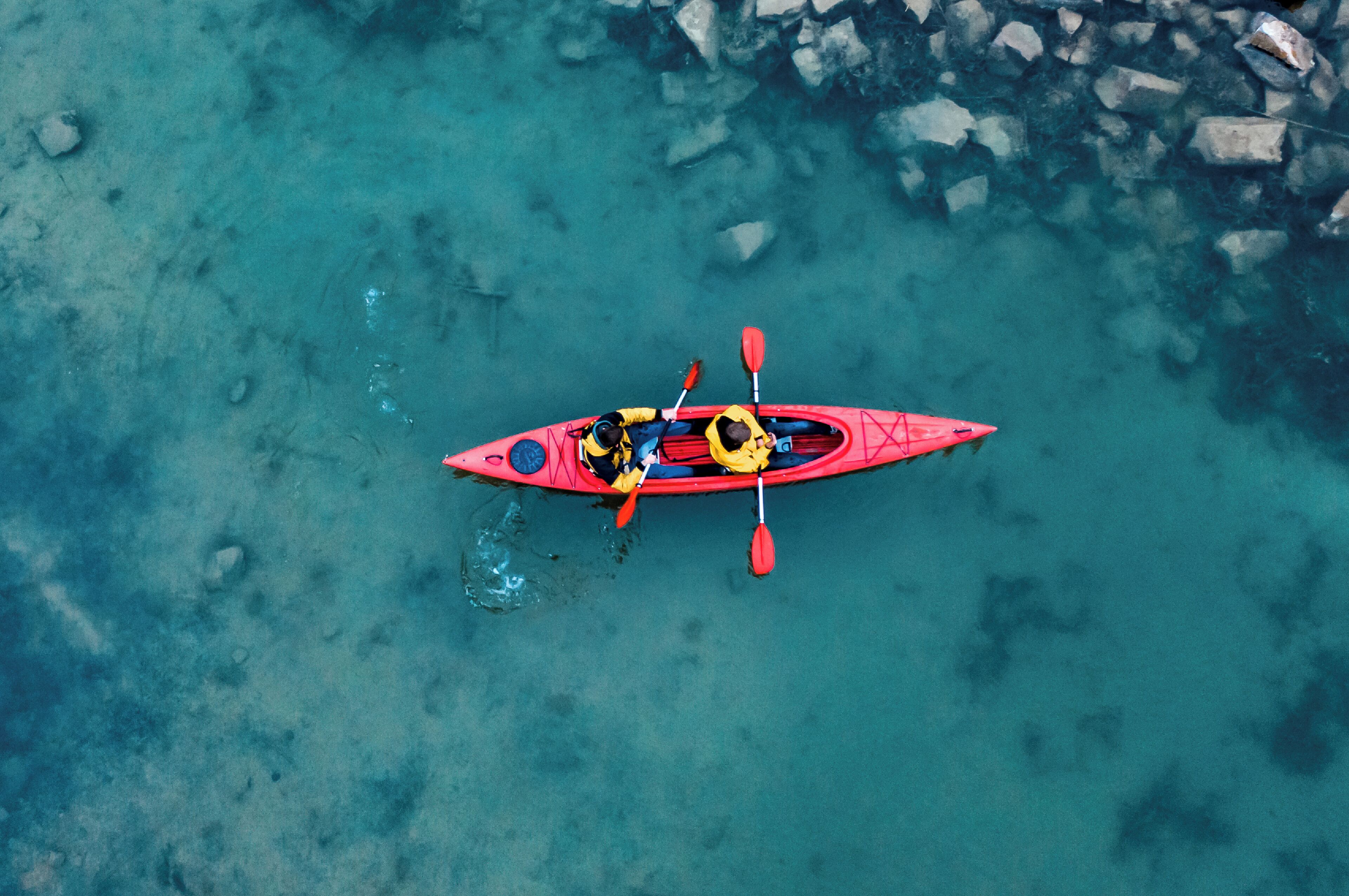 A red kayak with two paddlers on a clear turquoise water body with visible rocks.