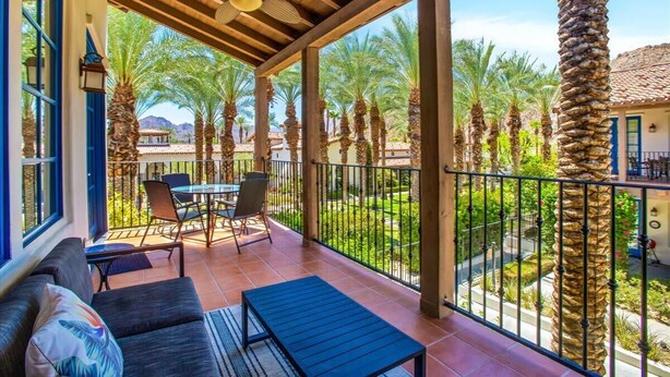 A covered outdoor area with a patio, seating, and a view of palm trees and a building in the background. - Hidden Palms A | 1BR Legacy Villa (La Quinta)