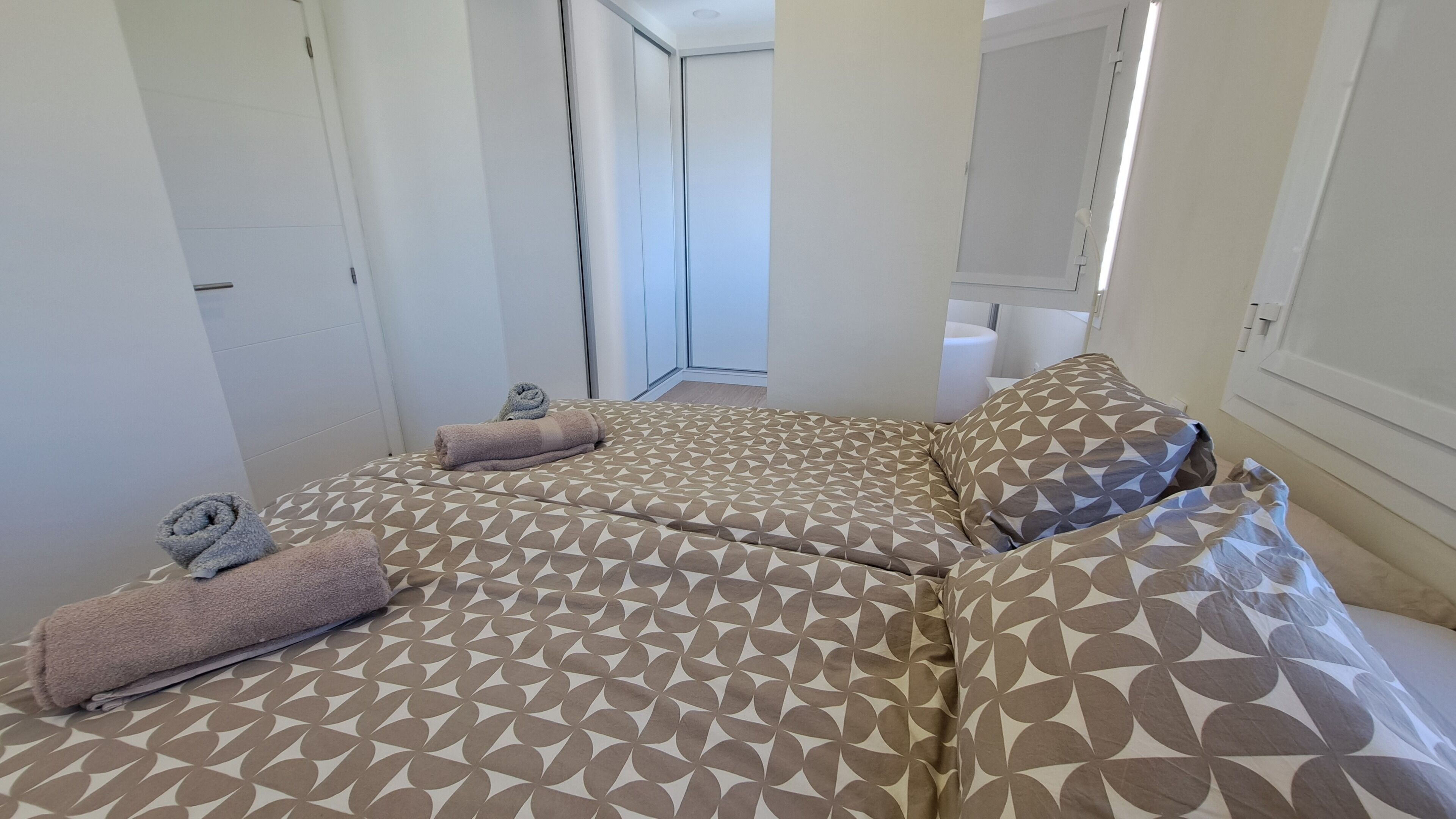 A hotel room with a bed featuring a geometric patterned bedspread, two rolled towels, and a pillow with a similar pattern.