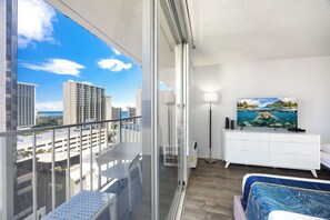 A balcony with a city view, a bed with a patterned bedspread, a white dresser with a painting, and a floor lamp.