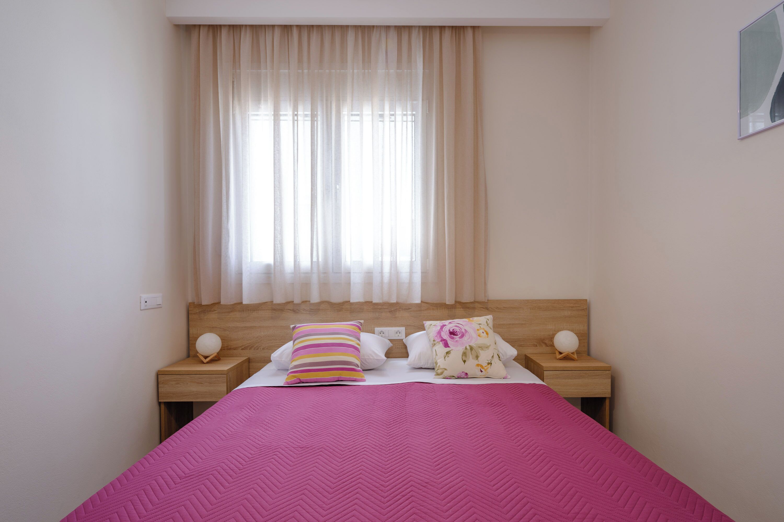 Una cama bien hecha con una colcha rosa, dos mesitas de noche de madera y una ventana con cortinas traslúcidas.