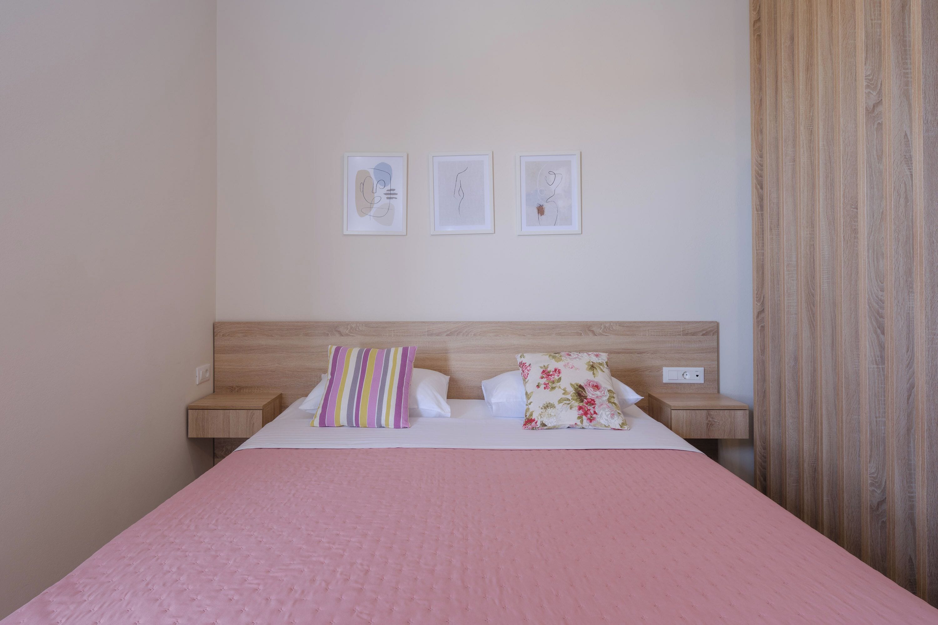 Ein ordentlich bezogenes Bett mit einer rosa Bettdecke, zwei Kissen und einem hölzernen Kopfteil. Darüber an der Wand hängen drei gerahmte Kunstwerke.