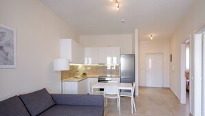 Een moderne woonkamer met een grijze bank, een eethoek met een witte tafel en stoelen, en een keuken met witte kasten en apparatuur.