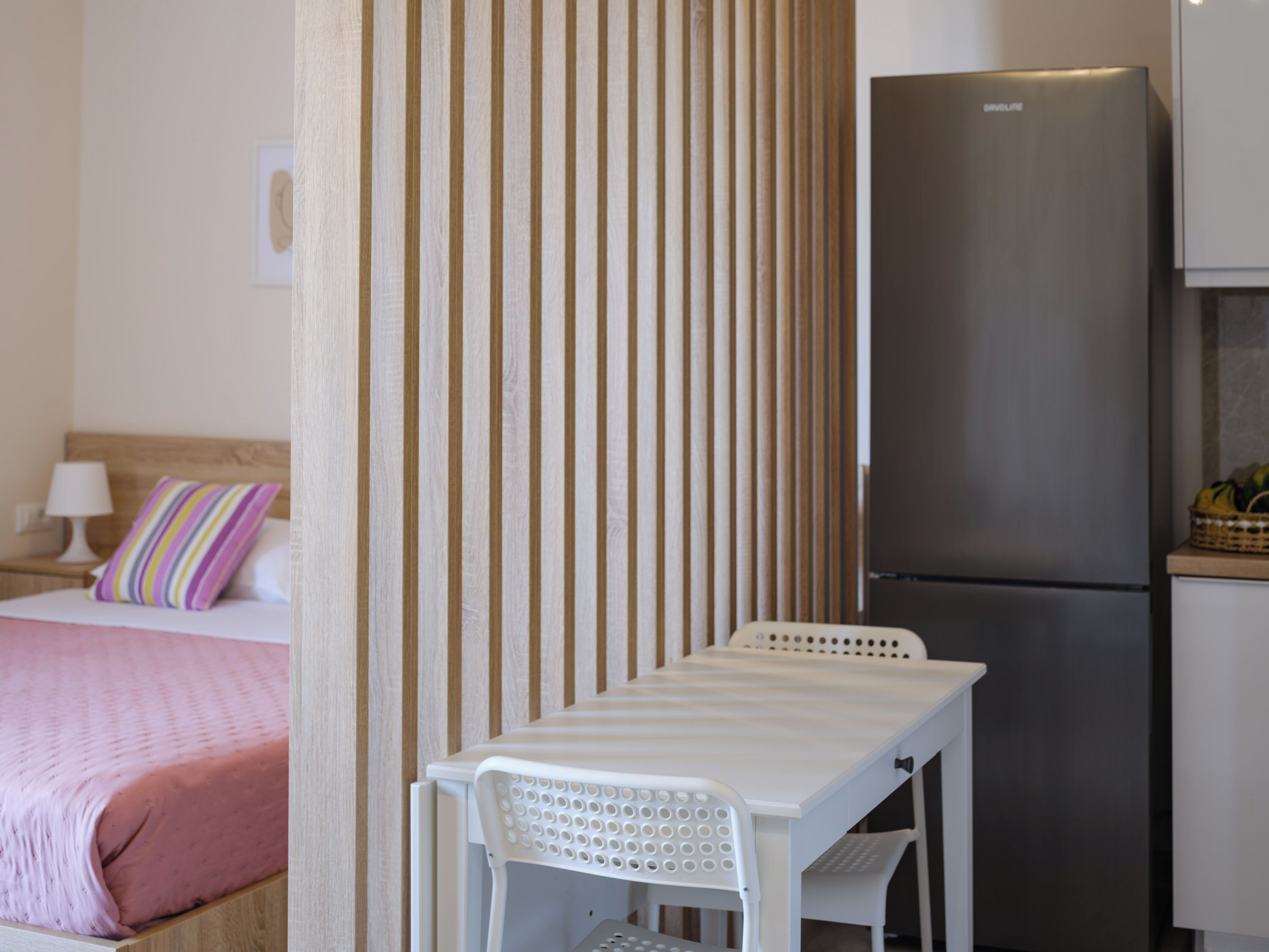 A room with a bed, a table with a chair, a refrigerator, and a wooden partition.