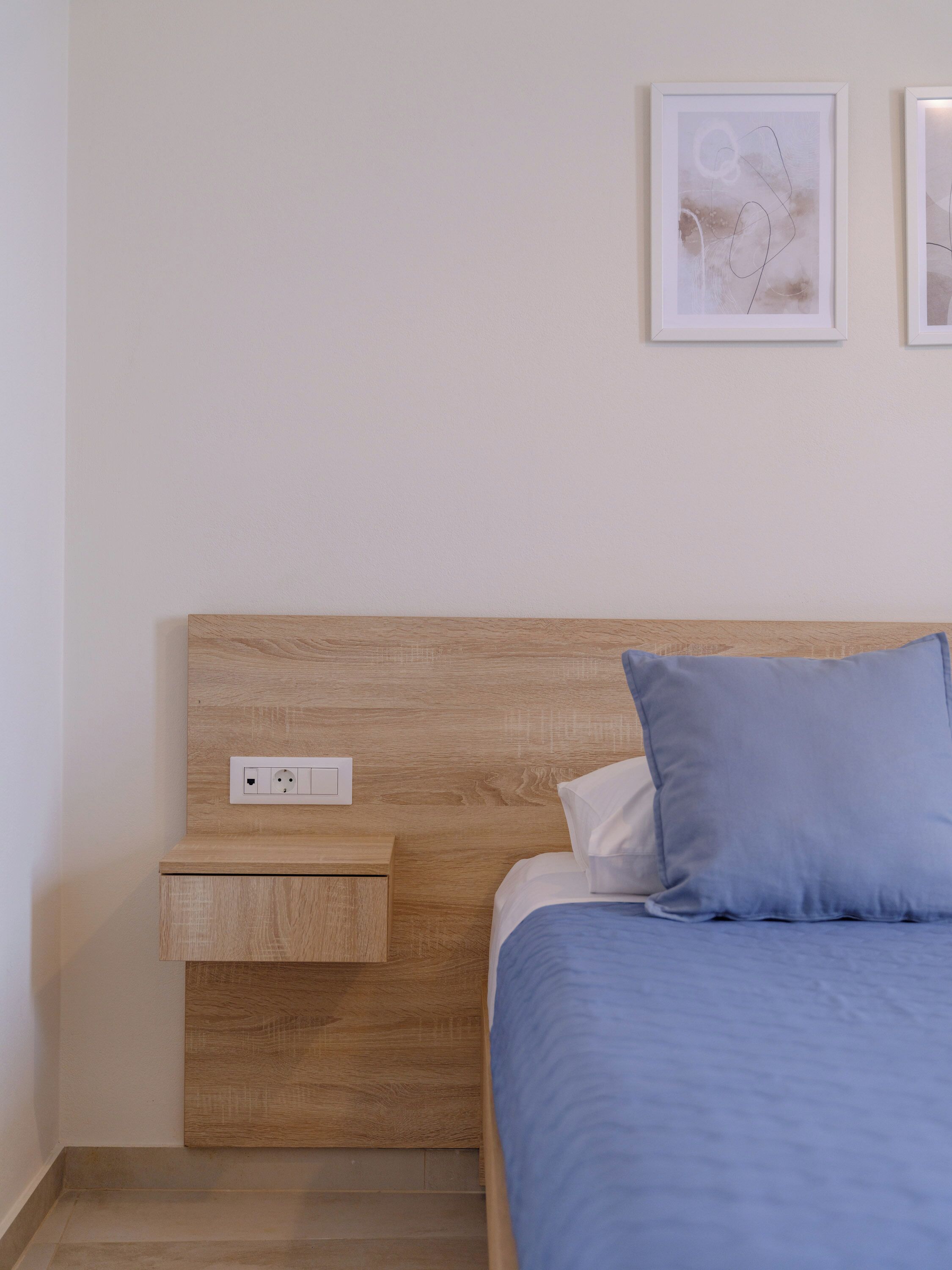 A bedroom with a wooden headboard, a bedside table, a bed with a blue pillow, and two framed abstract artworks on the wall.